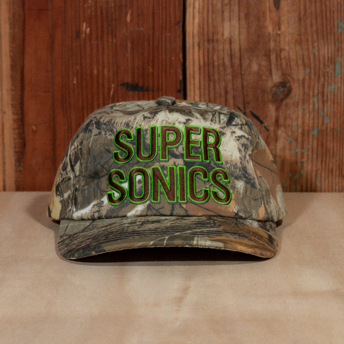 Peak Sonics Hat front view: woodland camo snapback with raised Super Sonics embroidery, on a wooden backdrop