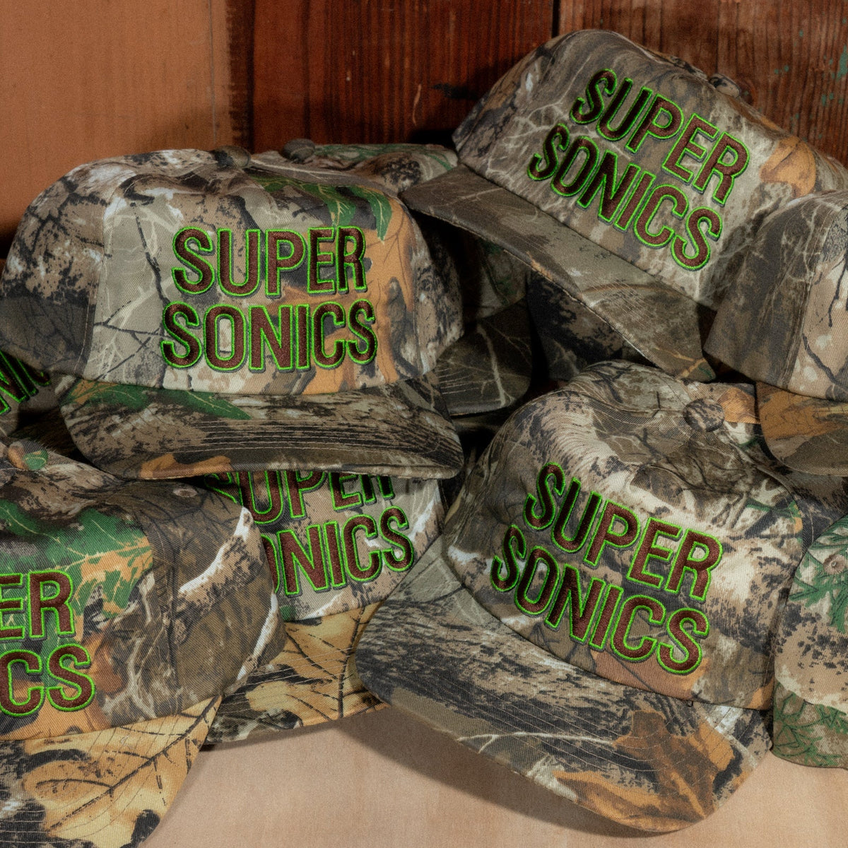 A pile of Peak Sonics Hats in woodland camo, with Super Sonics embroidery facing every direction