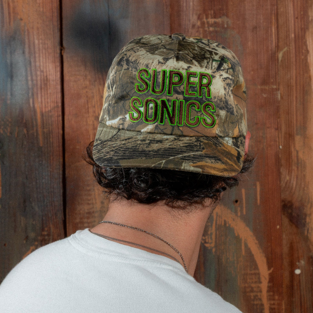 Model in a white T-shirt wearing the Peak Sonics Hat, head tilted down to show the embroidery