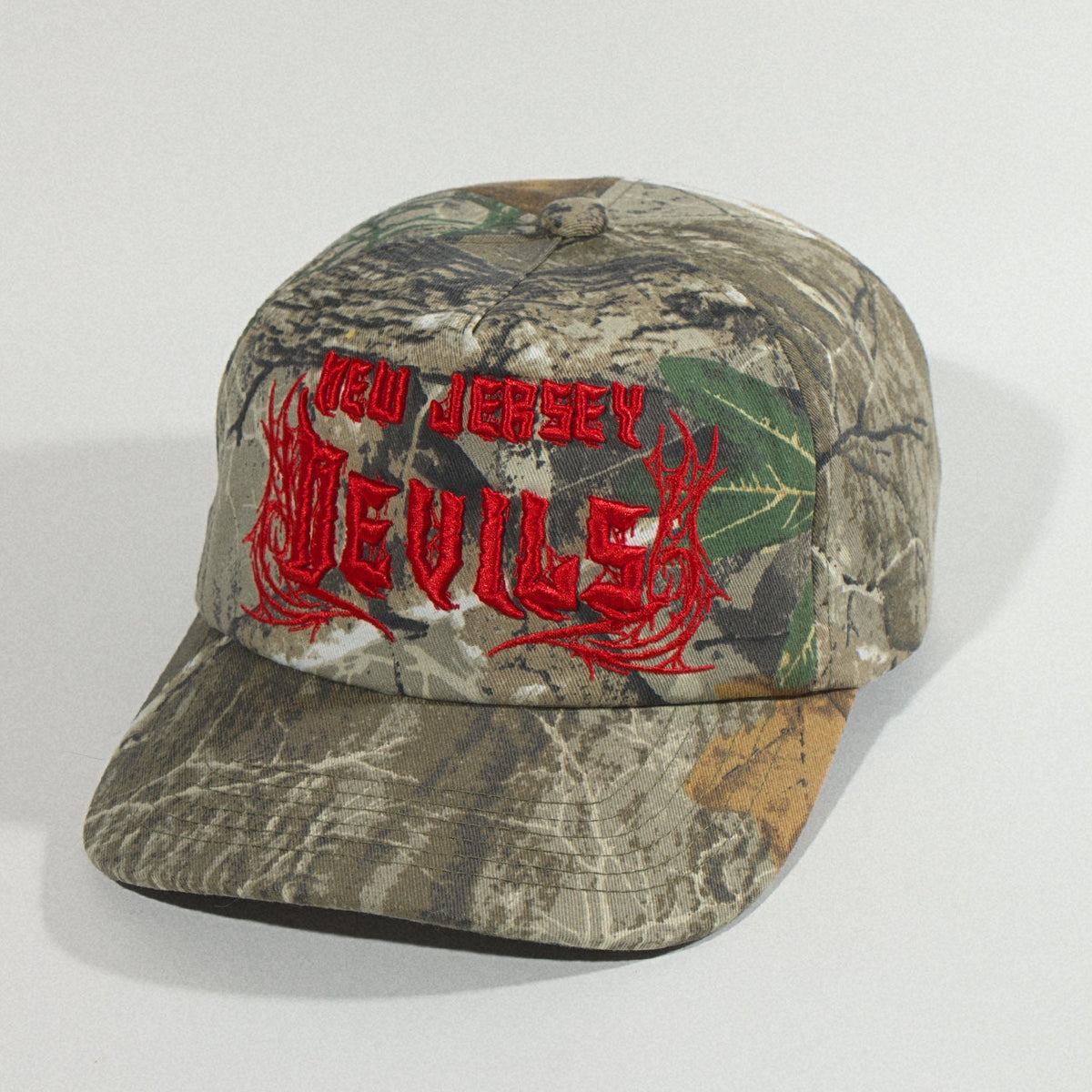 Three-quarter view from the left showing the red embroidered Devils logo and the curved camo brim